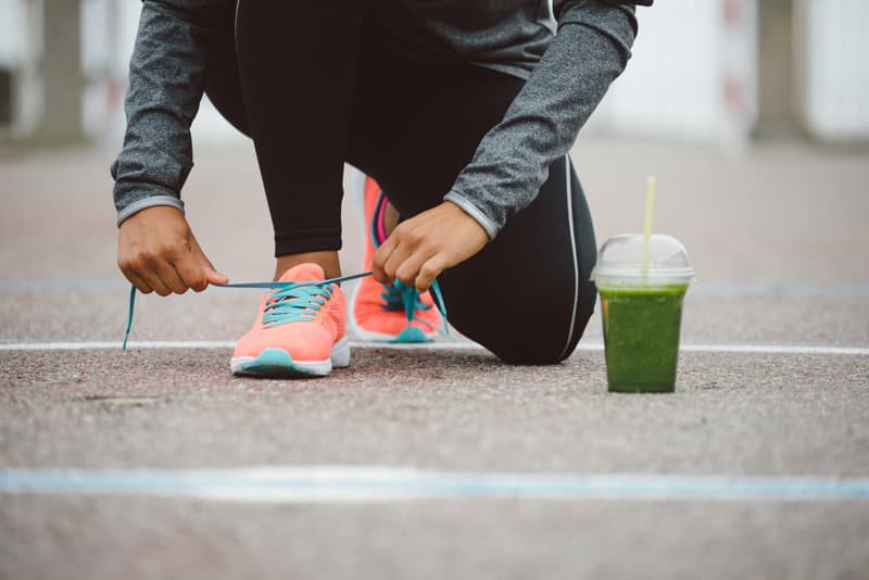 Close-up of a runner tying shoelaces on a track with a green smoothie nearby, emphasizing sports nutrition for endurance training.