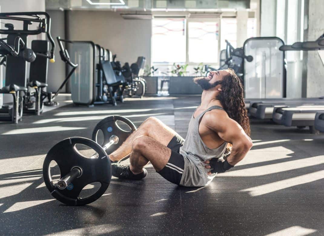 Portrait of injury young adult man athlete with long curly hair working out in gym, sitting on floor and have strong hurt problem with back, spasm painful. Gripping spinal with hand, screaming. Indoor