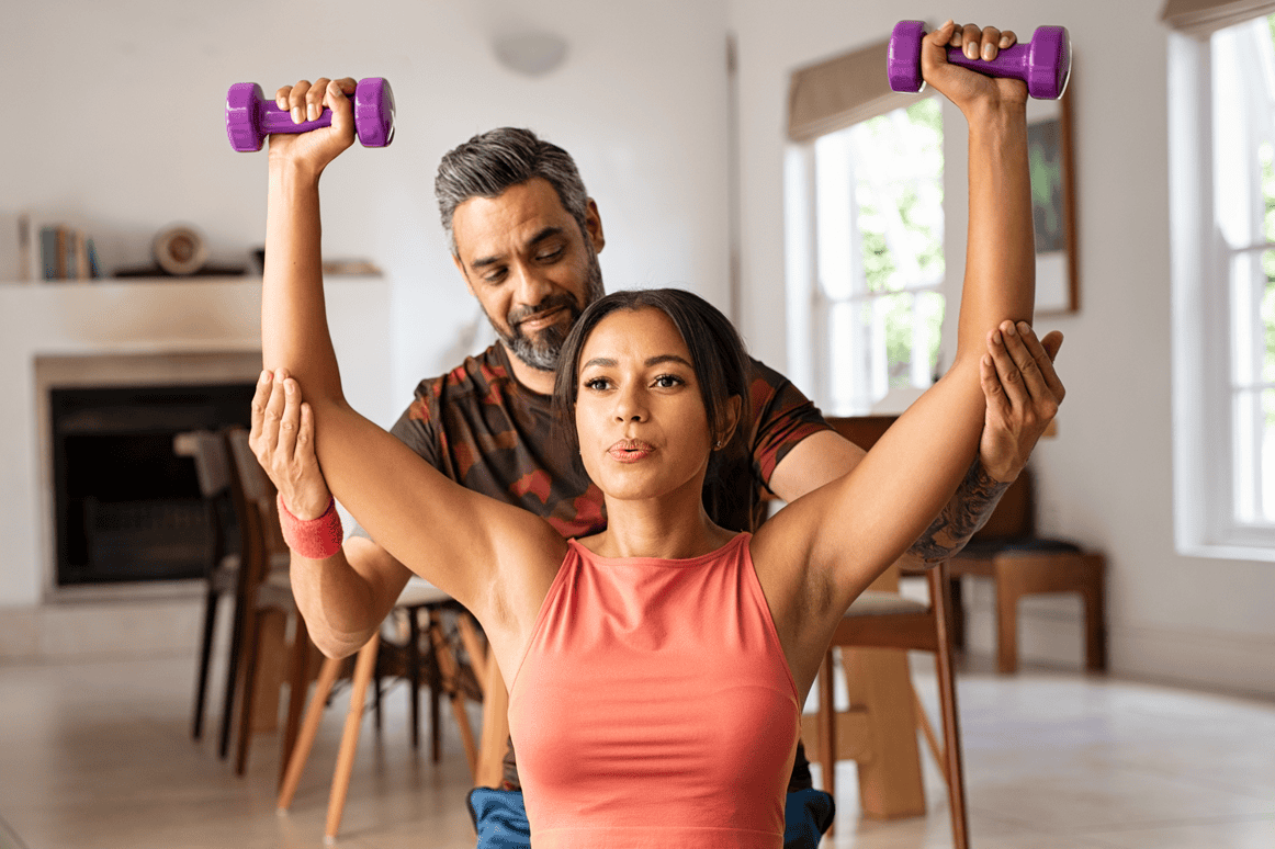 Sports rehabilitation specialist assisting woman with shoulder exercises using light dumbbells for rotator cuff strengthening. Focus on shoulder stability and injury prevention.