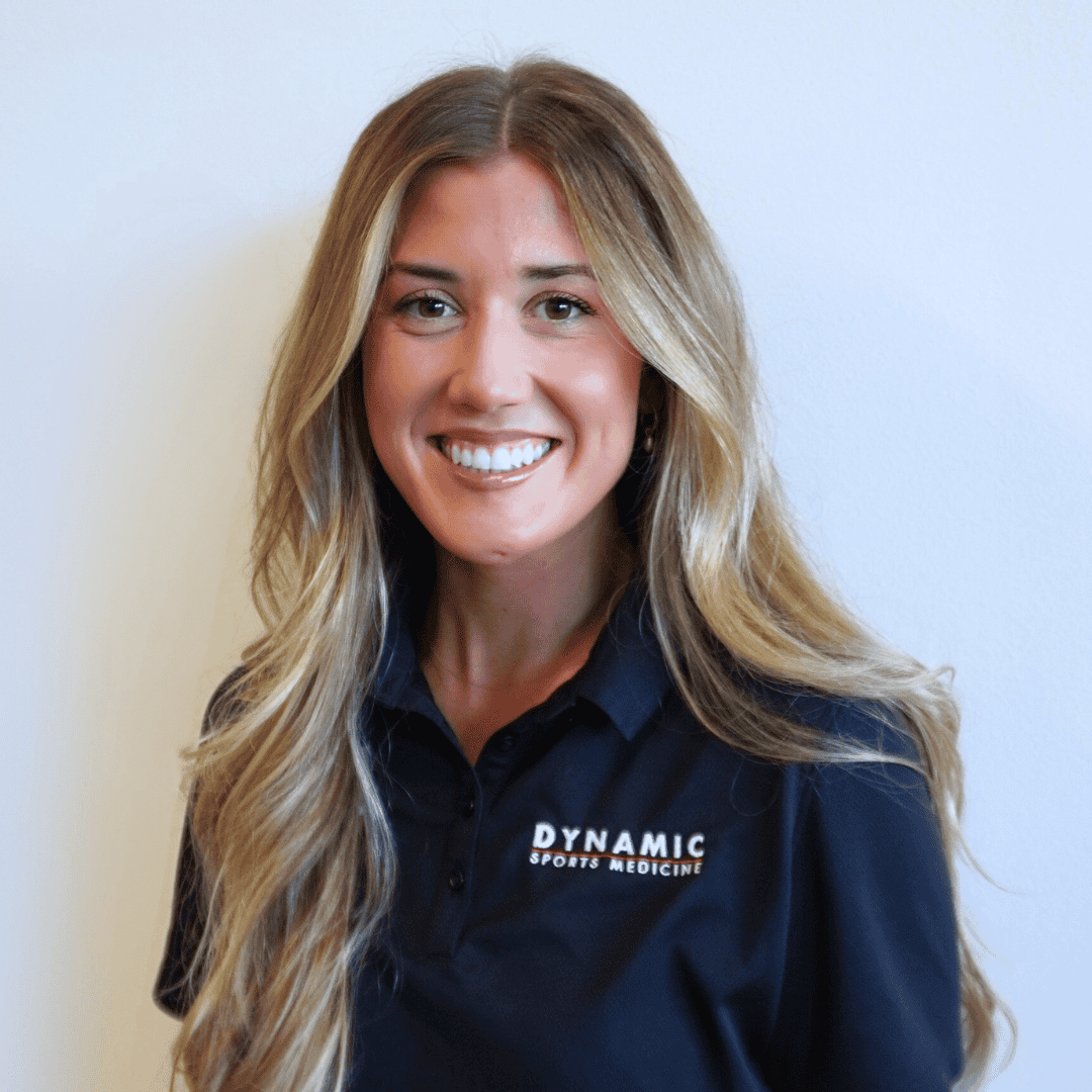 Athletic woman smiling in sports medicine office, wearing a black polo shirt with "Dynamic Sports Medicine" logo, emphasizing expert sports injury care and injury prevention services.