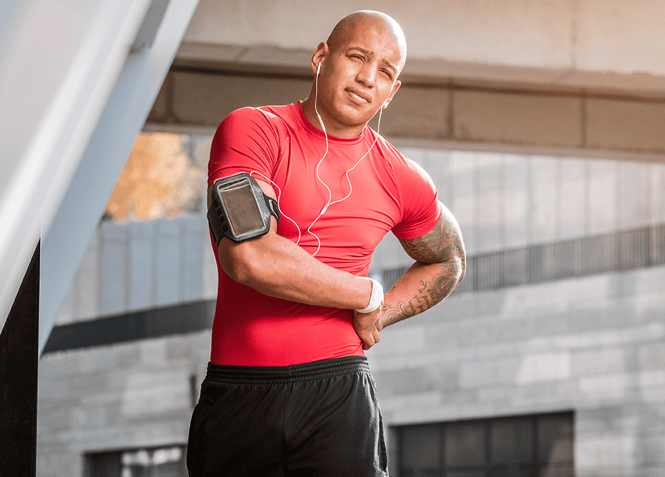 Active man with sports injury discomfort, holding his side after exercising, wearing athletic gear, and listening to music with earphones at a sports facility.