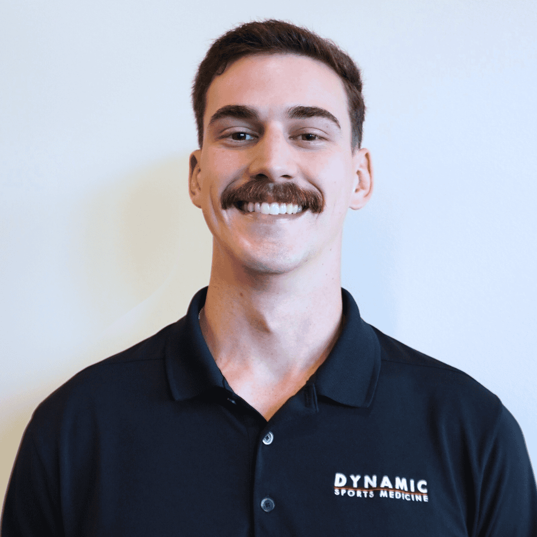 A smiling male sports medicine professional in a black polo shirt with "Dynamic Sports Medicine" logo, promoting expert injury care and athletic performance recovery services.