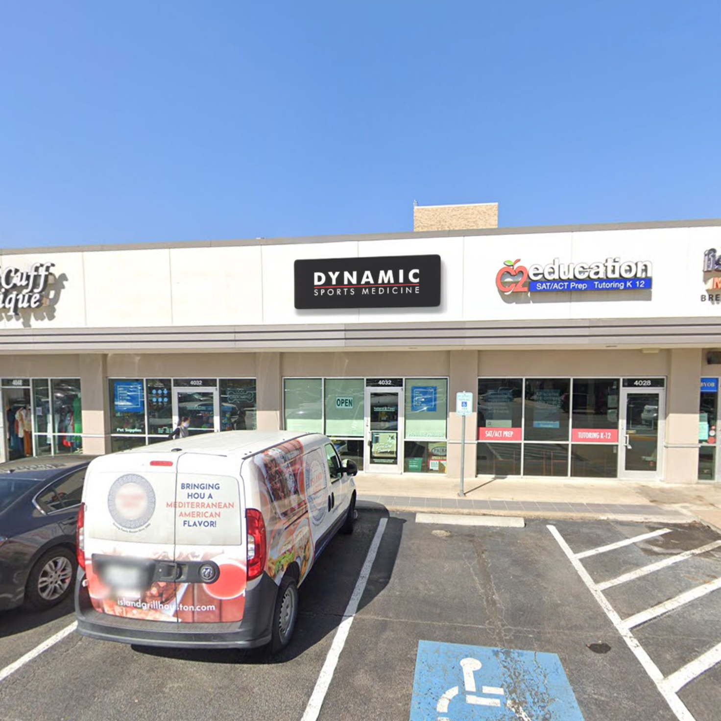 Exterior view of the Dynamic Sports Medicine clinic storefront with parking lot in front, showcasing the clinic's signage and surrounding retail stores.