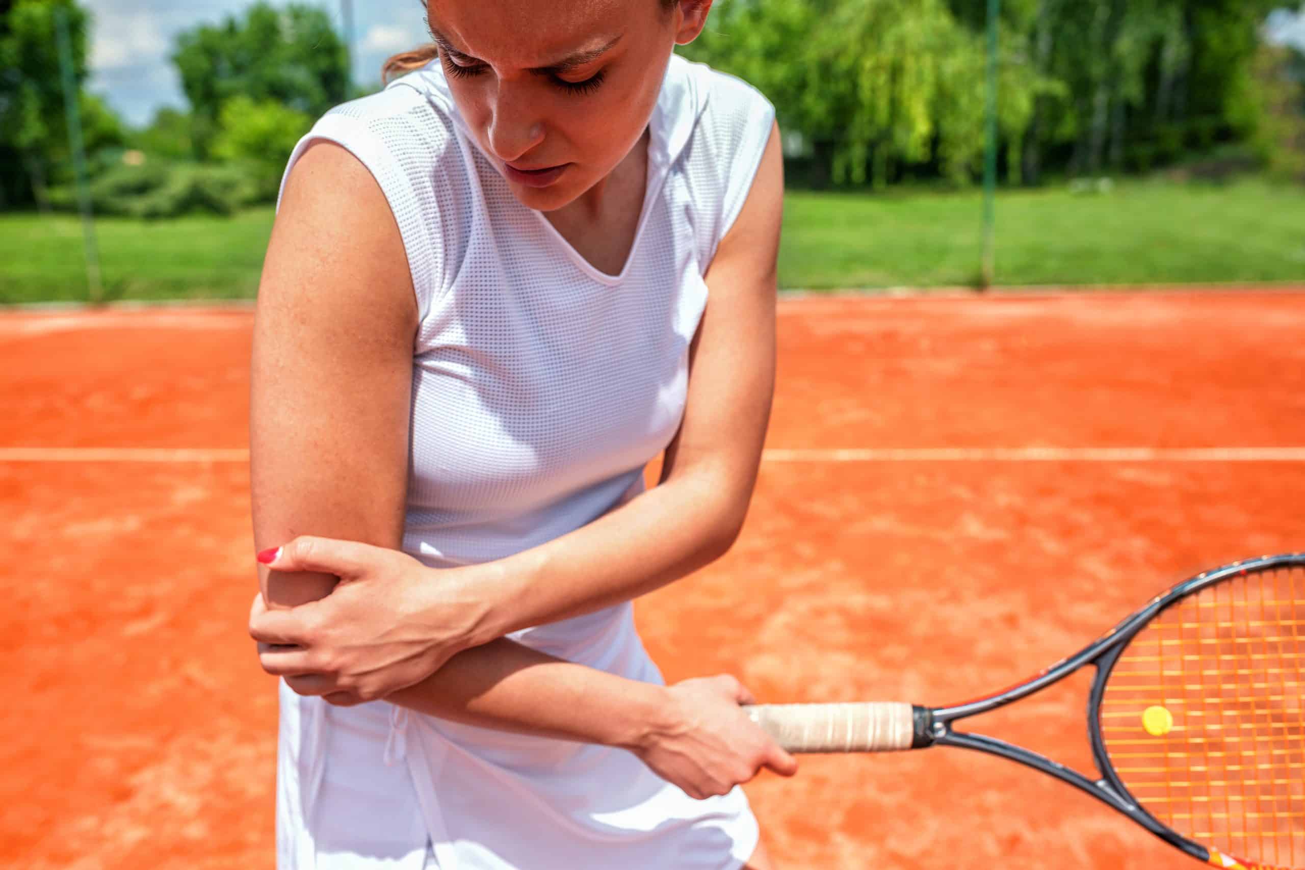 Female tennis player experiencing elbow pain during a match on an outdoor clay court, highlighting tennis elbow injury and the need for sports medicine treatment.