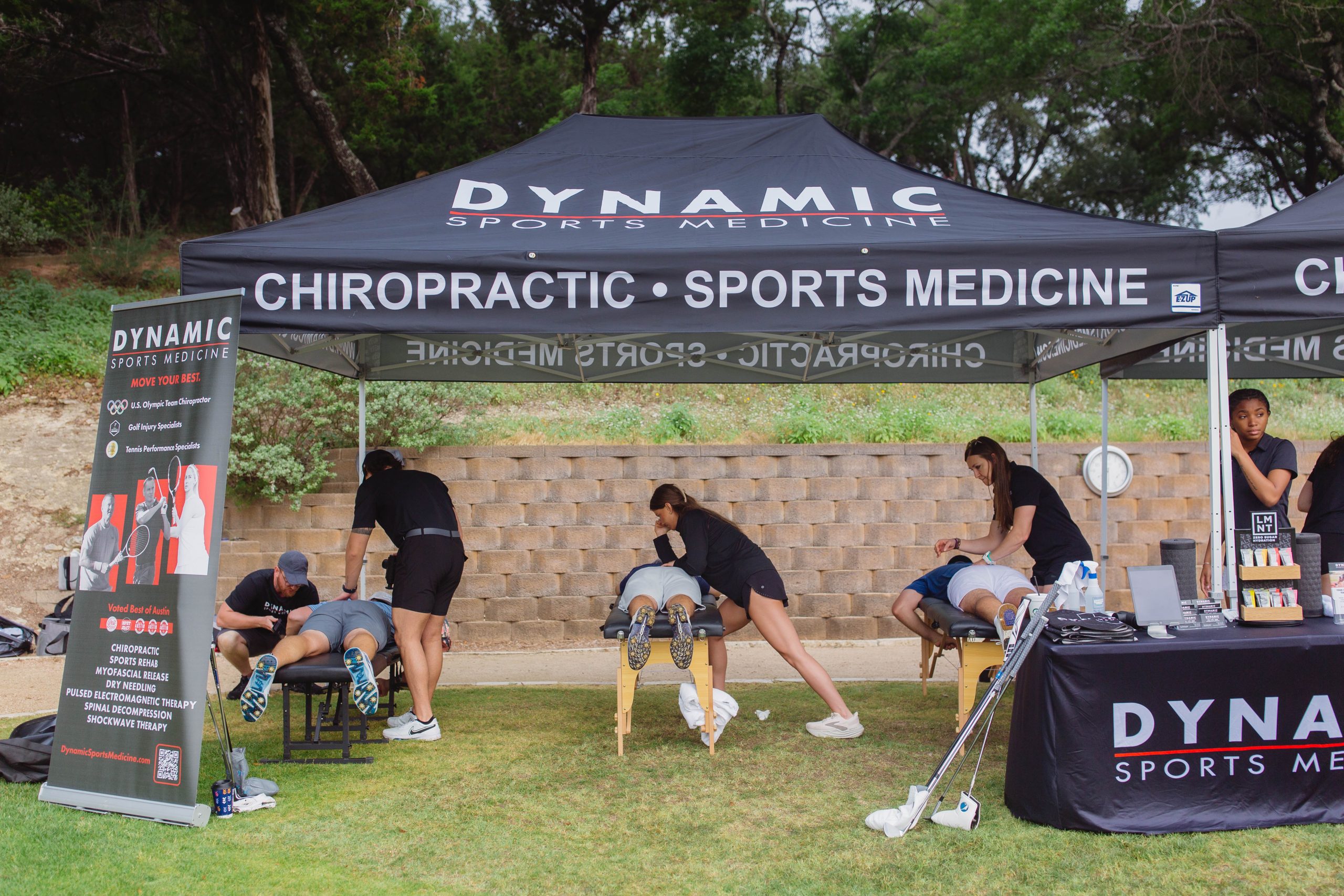 The image depicts a professional outdoor sports medicine and chiropractic care setup by Dynamic Sports Medicine. Several athletes are lying on treatment tables while therapists provide hands-on therapy. The scene is set in a grassy area with trees in the background, under a branded tent and informational banner, emphasizing expert sports injury management and recovery services.