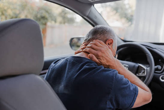 Man sitting in the driver's seat of a car, gripping the back of his neck in pain after an accident.