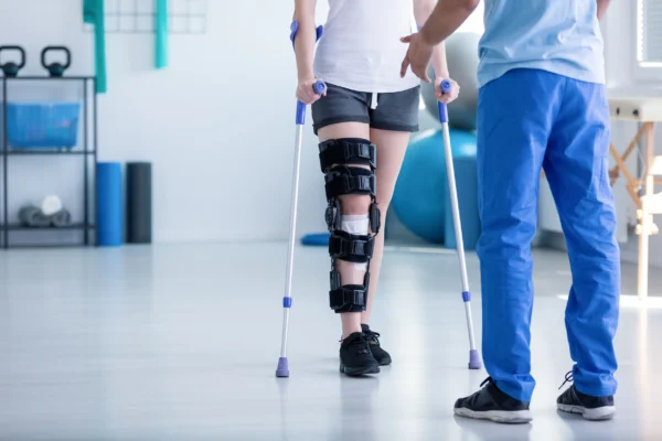 Patient in a knee brace using crutches while walking with a physical therapist in a rehabilitation gym.