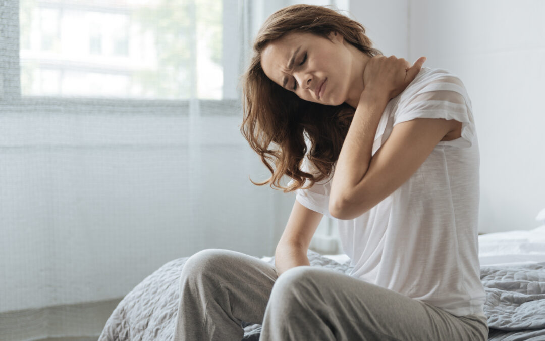 A young woman with long, wavy auburn hair sits on the edge of a bed, dressed in a white t-shirt and gray sweatpants in what appears to be a home setting. Her eyes are closed and her expression shows clear discomfort as she reaches back with one hand to hold her neck and shoulder. Her shoulders curl forward, and her whole posture reads as stiff and pained. Soft daylight filters through sheer curtains behind her, giving the scene a quiet, everyday, morning-at-home feel.