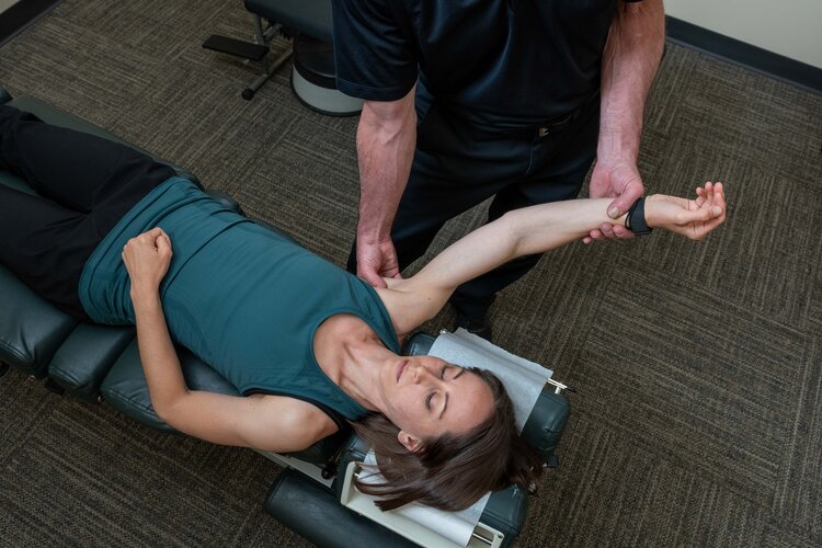This image shows a physical therapist performing Active Release Technique (ART) on a female patient lying face-up on a padded treatment table. The practitioner, dressed in all black, uses both hands to manipulate the patient's shoulder while extending her arm outward. The patient, wearing a teal top, has her eyes closed as the treatment is performed in a professional clinical setting.