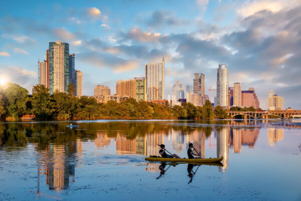 Two people paddling a yellow kayak across Lady Bird Lake at sunrise, with the Austin, Texas downtown skyline reflected in the calm water behind them.