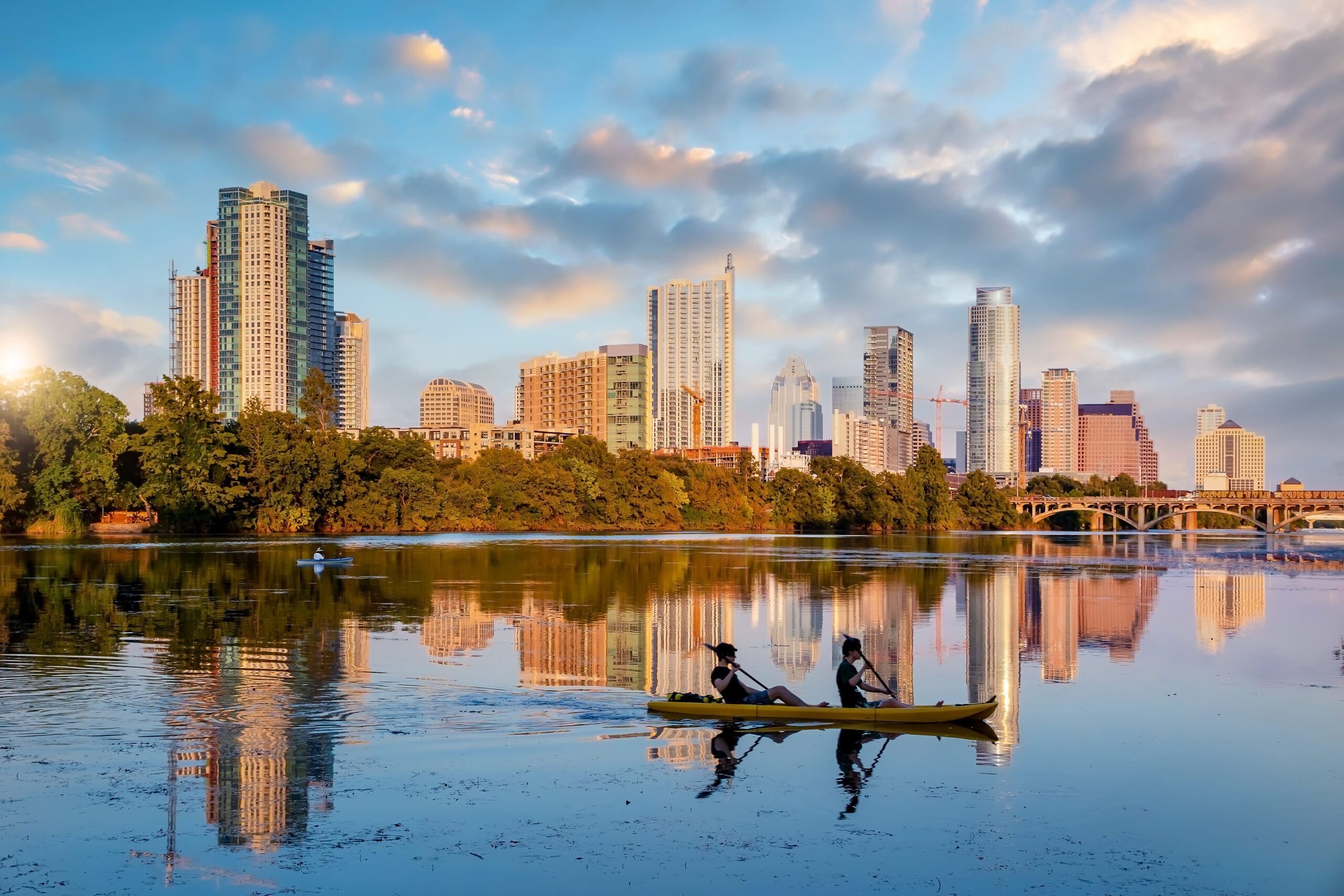 Golden-hour view of the Austin, Texas skyline reflected in Lady Bird Lake. A row of downtown high-rises catches warm early-morning light against a partly cloudy blue sky, with a tree-lined shoreline separating the buildings from the water. In the foreground, two people paddle a yellow tandem kayak/paddleboard across the still water, their silhouettes and paddle strokes mirrored perfectly in the glassy surface. A stone arch bridge and a lone paddleboarder are visible further back near the shoreline.