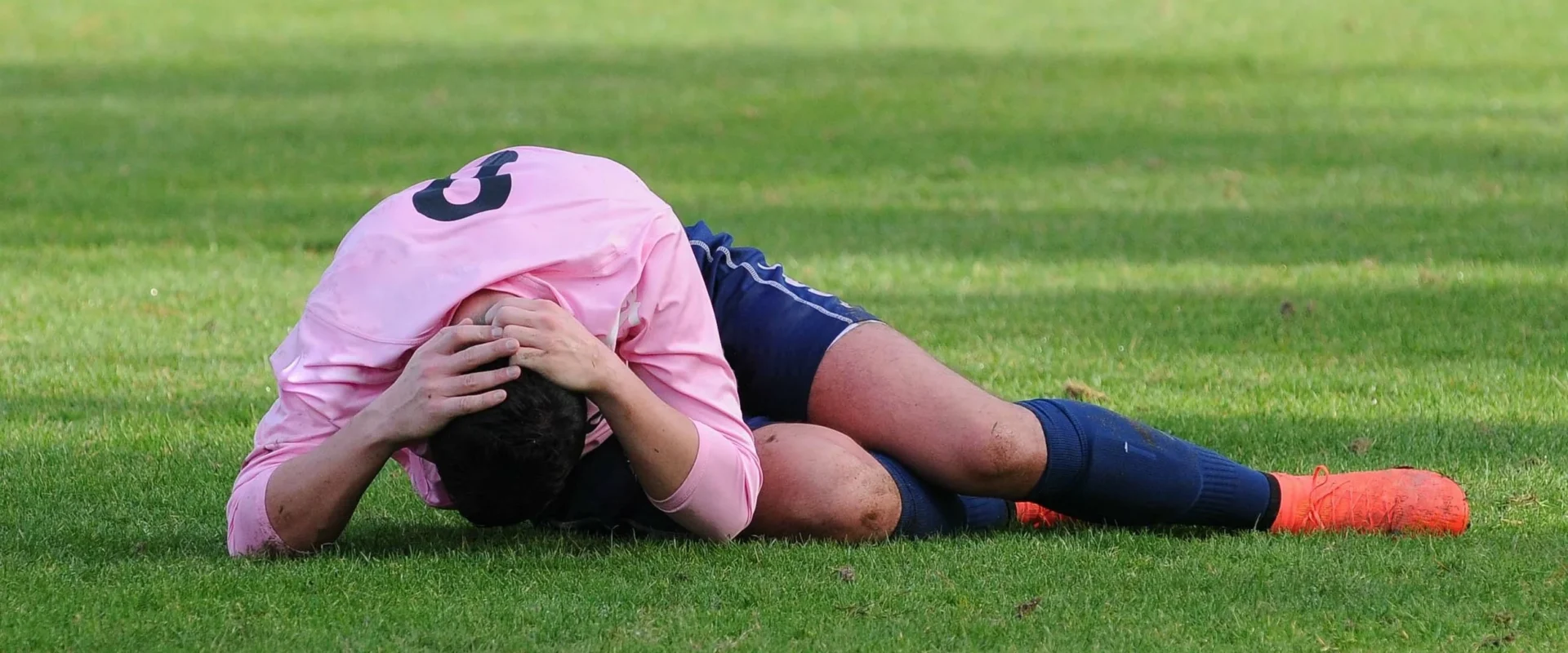 A soccer player in a pink jersey and navy shorts lies curled on the field, holding his head with both hands.