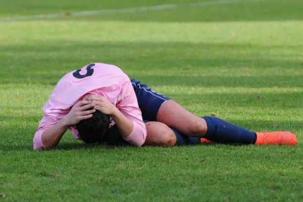 A soccer player in a pink jersey and navy shorts lies curled on the field, holding his head with both hands.