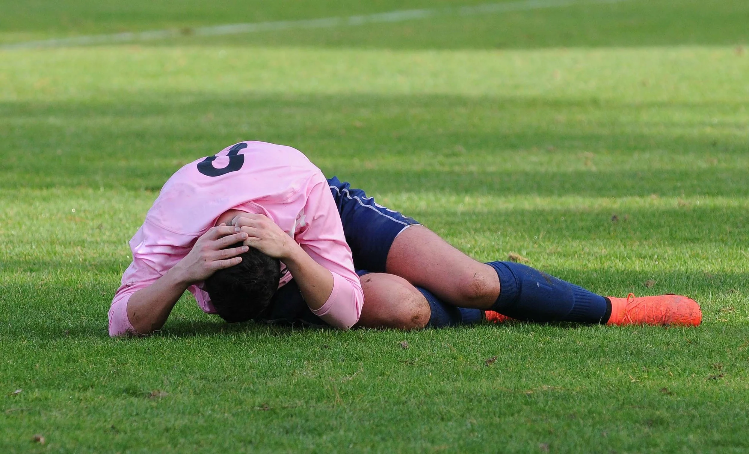 A soccer player lies curled on his side on a grass field, gripping his head with both hands after a fall or collision. He wears a pink number 5 jersey, navy shorts, navy socks, and orange cleats.
