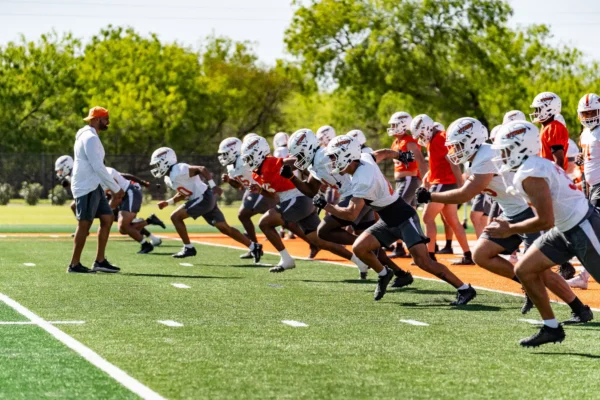 Football players in white and orange practice jerseys sprint off the line during a team conditioning drill on a turf field, with a coach in an orange cap looking on.