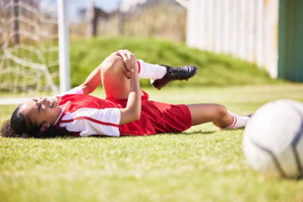 Young female soccer player lying on the field grimacing in pain while holding her knee, soccer ball in foreground.