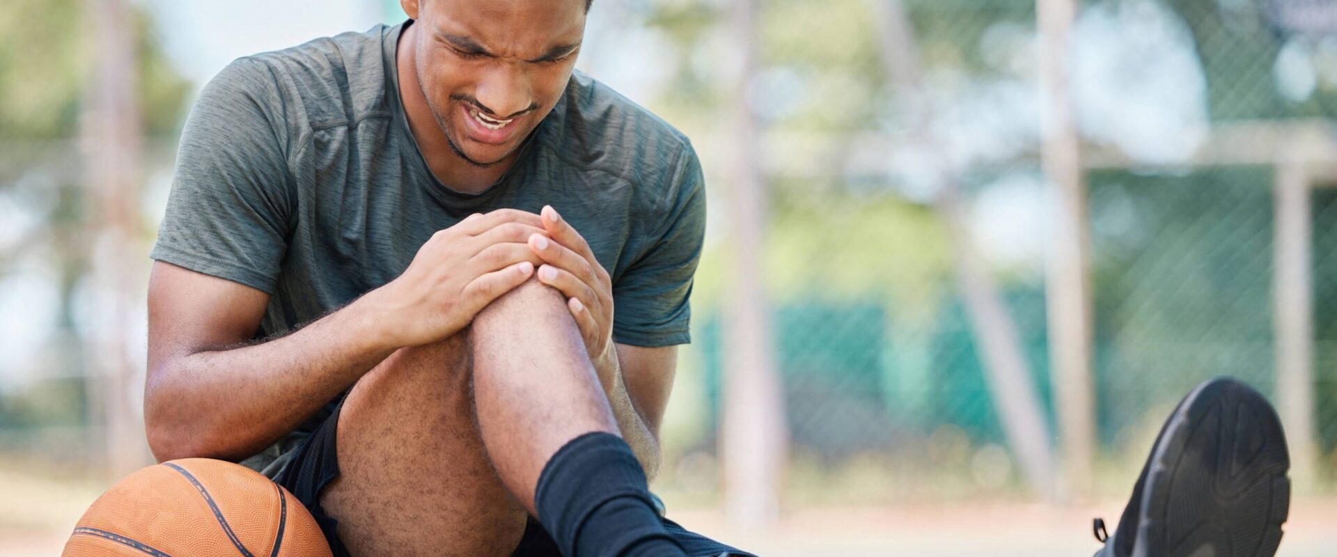 Basketball player sitting on an outdoor court holding his knee in pain, basketball beside him.
