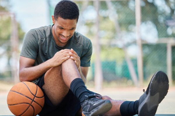 Basketball player sitting on an outdoor court holding his knee in pain, basketball beside him.