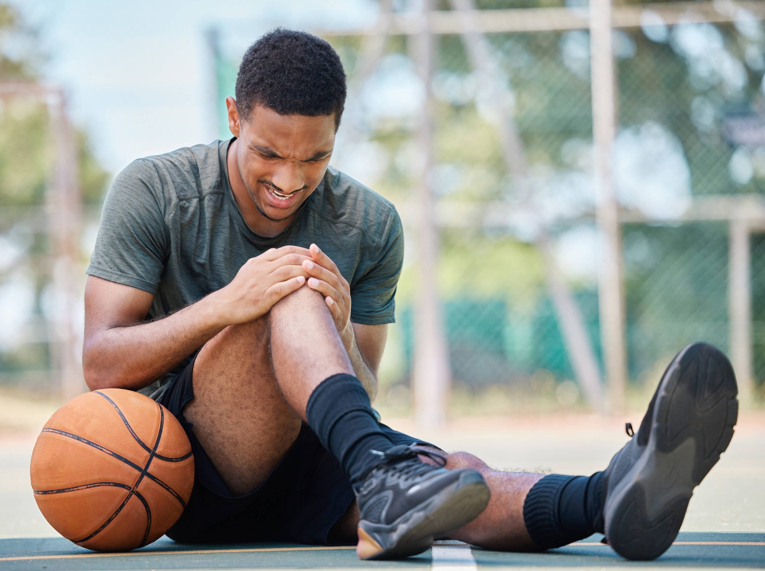 A young male basketball player sits on an outdoor court, grimacing and clutching his knee with both hands. His basketball rests beside him and his cleats/shoes are visible in the foreground, with a chain-link fence and trees blurred in the background. The image conveys an acute injury moment, suitable for content addressing knee pain, overuse injuries, or the importance of prompt sports medicine evaluation.