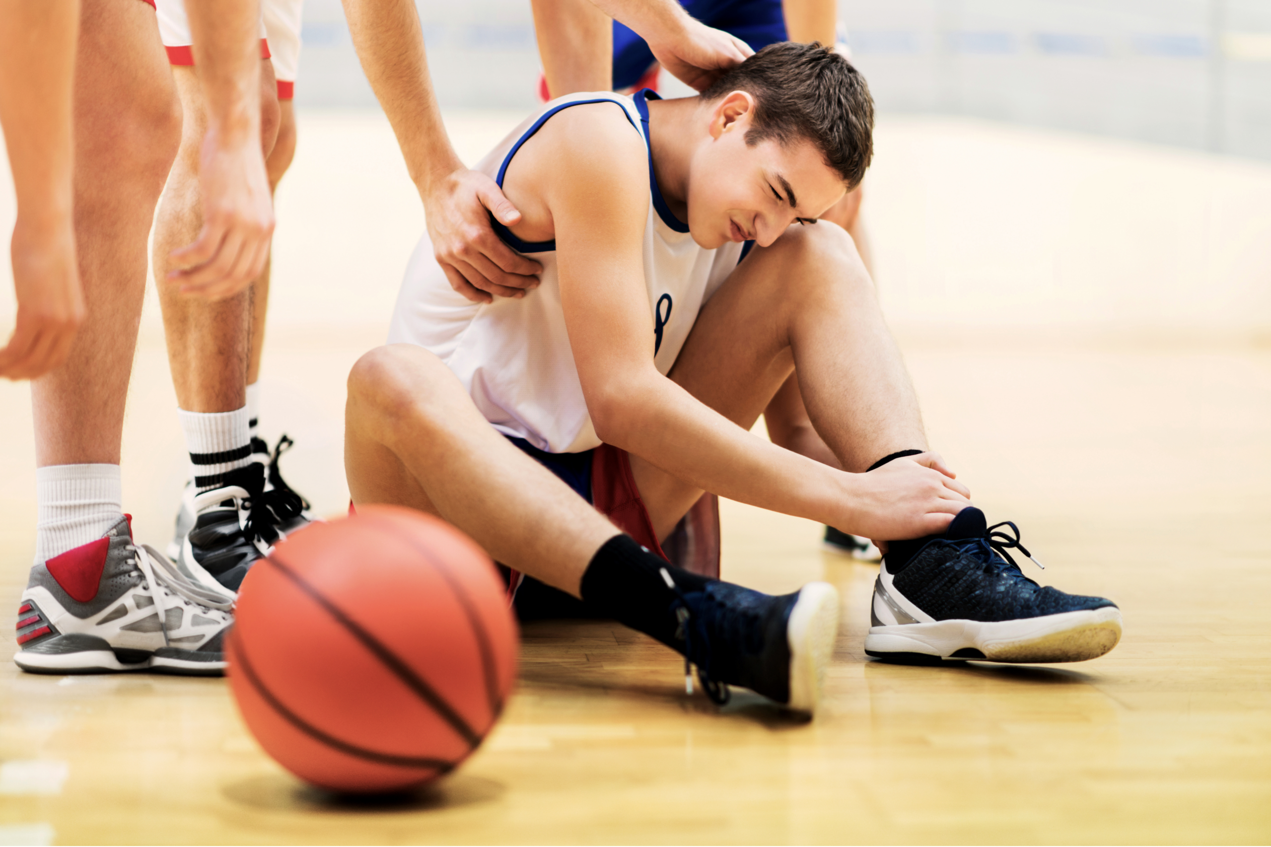 A teenage basketball player wearing a white and blue jersey sits on an indoor gym floor, wincing in pain and gripping his ankle with one hand while a teammate places a supportive hand on his shoulder. A basketball rests on the court in the foreground, and other players' legs are visible standing around him. The image captures the moment after an in-game injury, well-suited for content on youth sports injuries, ankle sprains, growth plate concerns, or the importance of proper medical evaluation for young athletes.