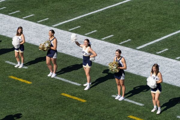 Five cheerleaders in navy and white uniforms with gold and white pom-poms stand spread out along the sideline of a sunny football field.