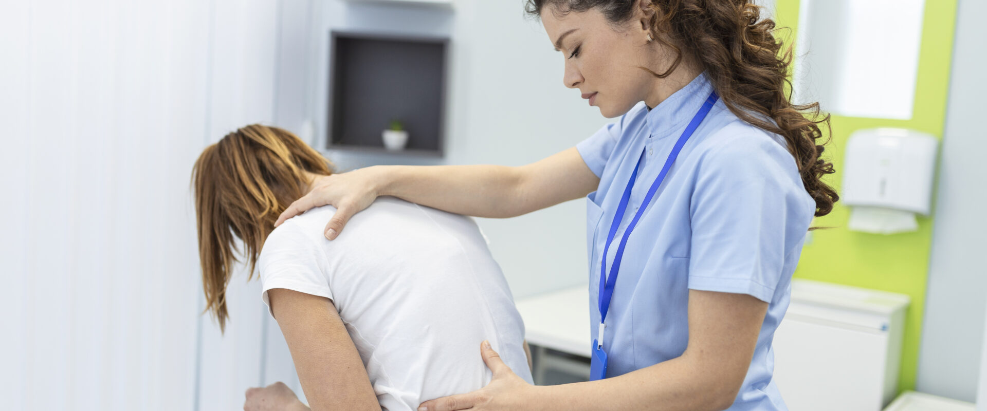 A healthcare provider in light blue scrubs places one hand on a seated patient’s shoulder and the other on her lower back while the patient leans forward on an exam table.