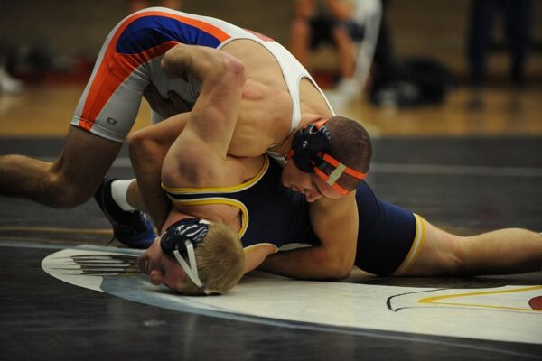 Two high school wrestlers in headgear grappling on a mat, one in a navy singlet controlling the other from above.